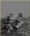 Inspecting and recording the remains of a fallen Japanese soldier on Attu. There appears to be a fencing mask and attire in the rubble.  [George Villasenor]