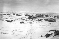 Freight dock right of center, dispensary in foreground. Generator shack center with ambulance parked left of shack. Foot prints in snow show path leading to our 6-man quarters. Steel mat runway between dispensary and freight dock. [Don Blumenthal]